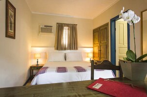 Een hotelkamer met een bed, nachtlampjes, een houten stoel, een tafel, een rode map, een plant in pot en een spiegel.