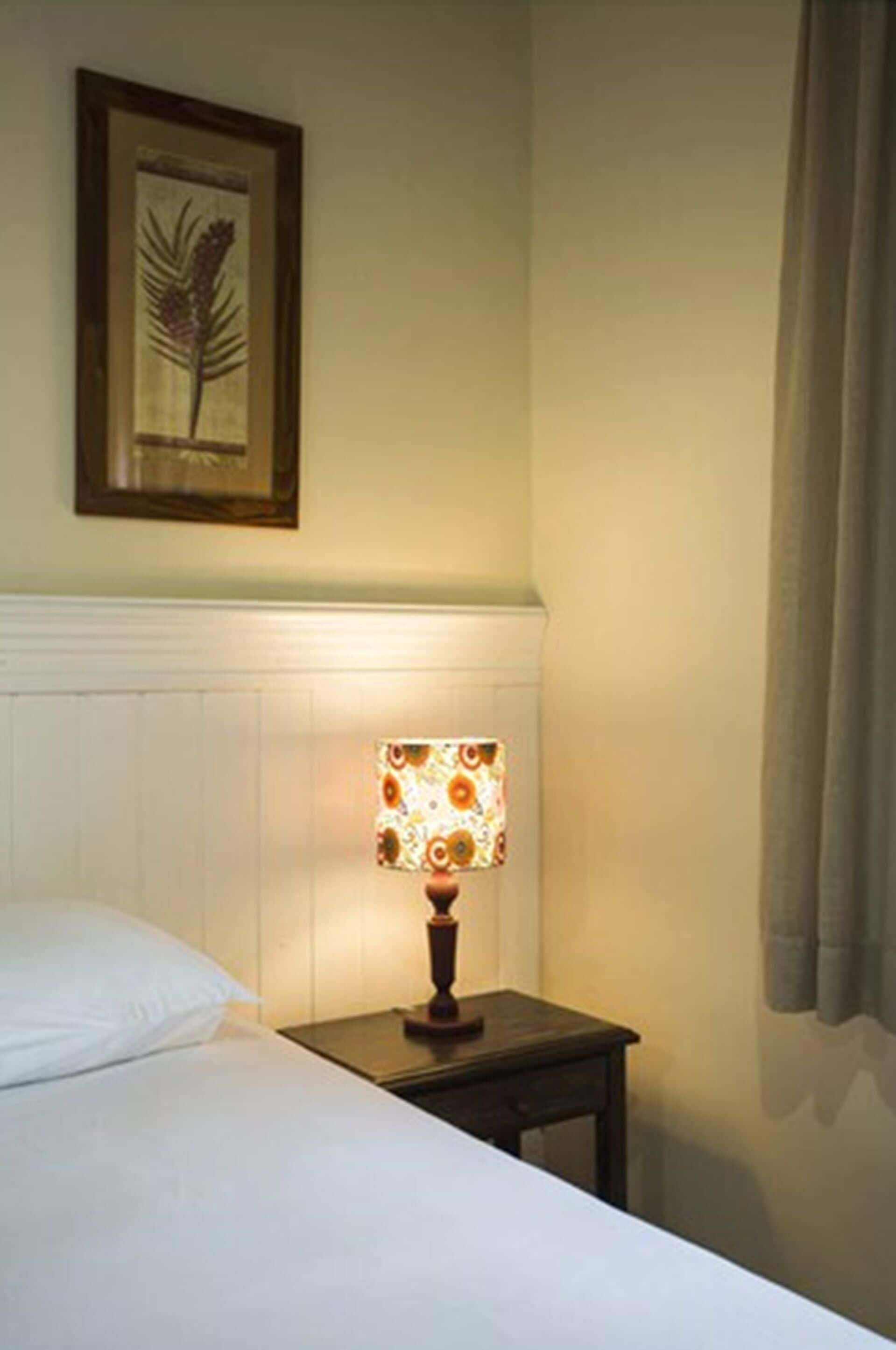A neatly made bed with white linens, a small bedside table with a lamp, and a framed botanical print on the wall.