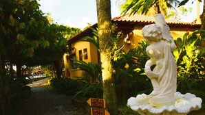 A statue on a pedestal in a garden with a building in the background. There are signs pointing to 'SALÃO DE CAFE,' 'ARTES,' and 'PISCINA E BAR.' - Pousada Corsário Paraty (Paraty)