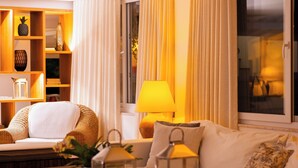 Una sala de estar acogedora con un sofá, una mesita de centro con velas y una lámpara. Se aprecia una estantería con objetos decorativos y una ventana con cortinas.