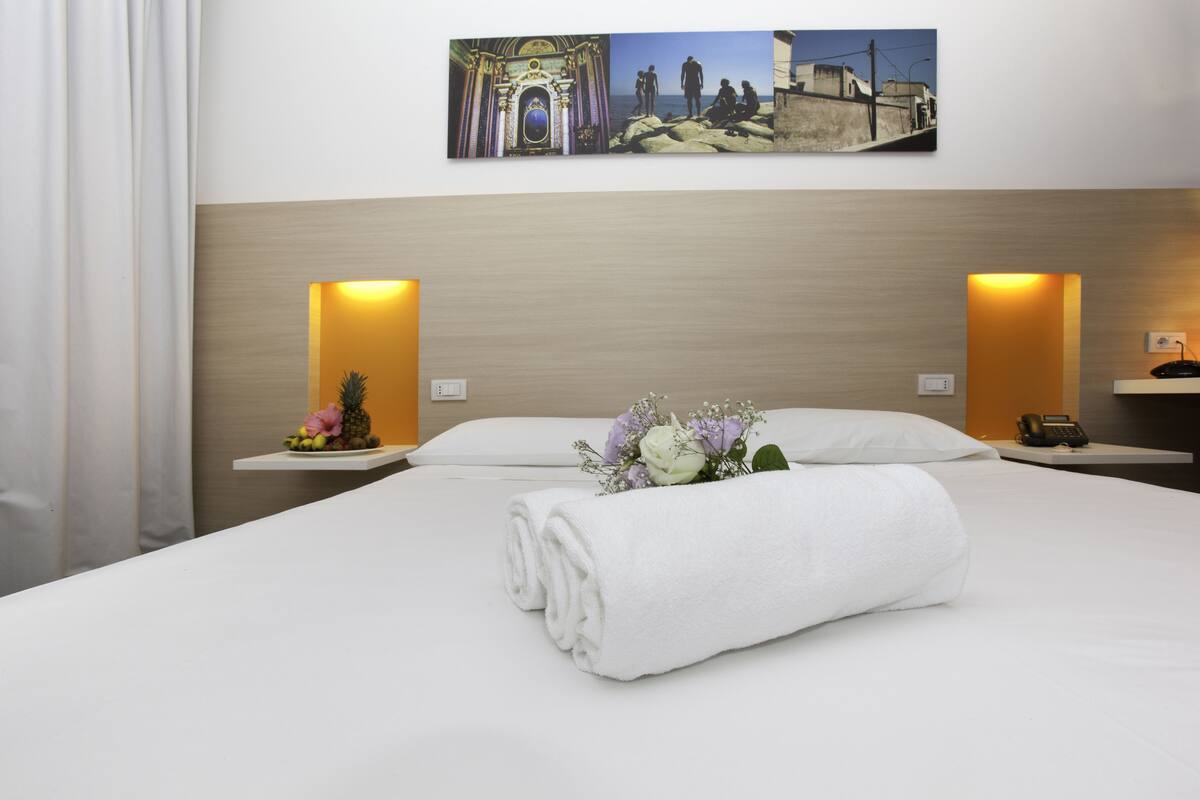 A hotel room with a neatly made bed, a wall-mounted artwork depicting a historical scene, and two bedside lamps.