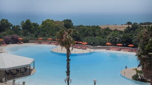 A large, clear swimming pool with red umbrellas and lounge chairs, surrounded by greenery and a view of the sea. - iH Hotels Agrigento Kaos Resort (Agrigento)