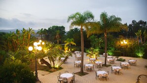 An outdoor dining area with tables and chairs, surrounded by palm trees and greenery. - iH Hotels Agrigento Kaos Resort (Agrigento)