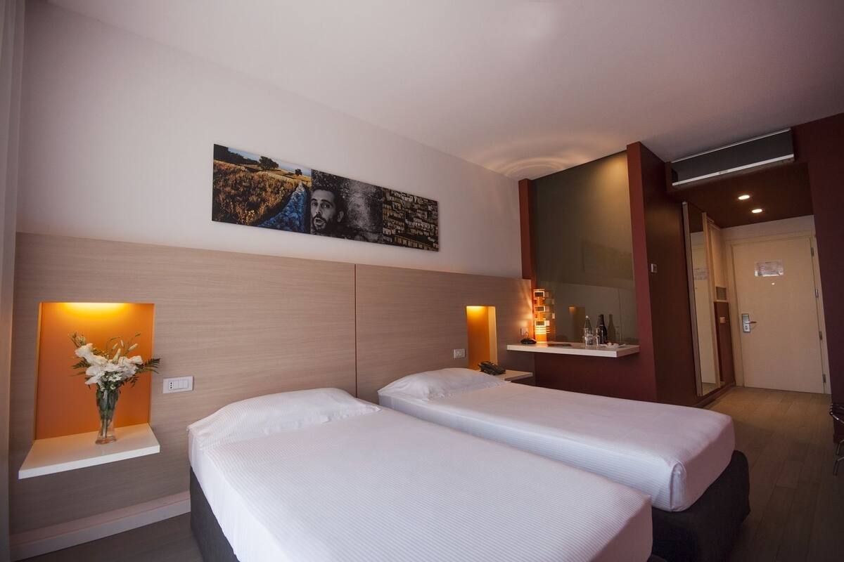 A hotel room with two beds, a large mirror, a wall-mounted artwork, and a bedside table with a vase of flowers.