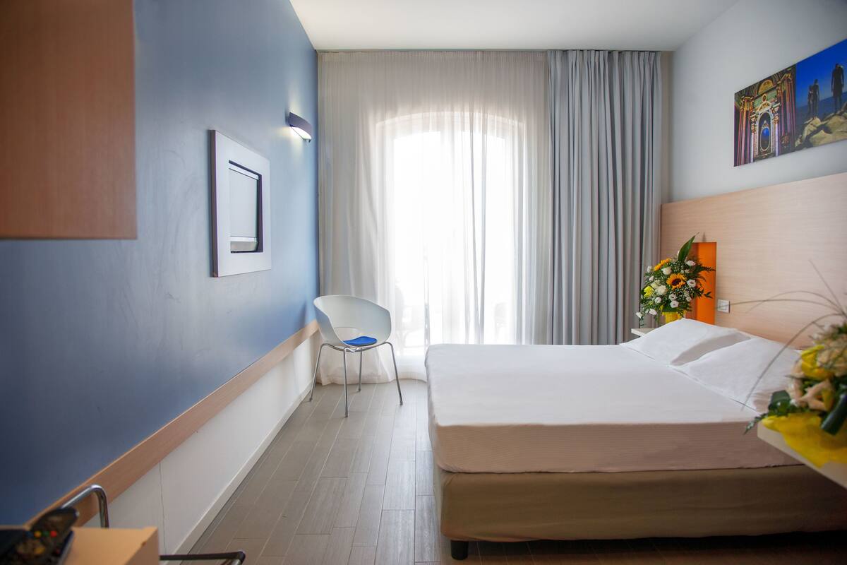 A hotel room with a bed, a chair, a desk with a vase of flowers, and a painting on the wall.