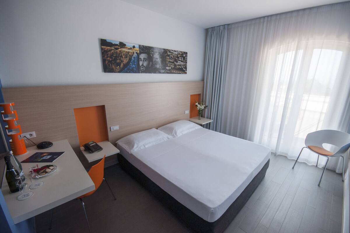 A hotel room with a bed, a desk with a chair, a telephone, and a painting on the wall.