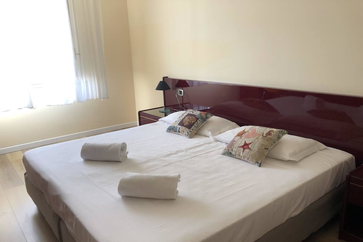 A neatly made bed with white linens and decorative pillows, a wooden headboard, and a bedside table with a lamp.