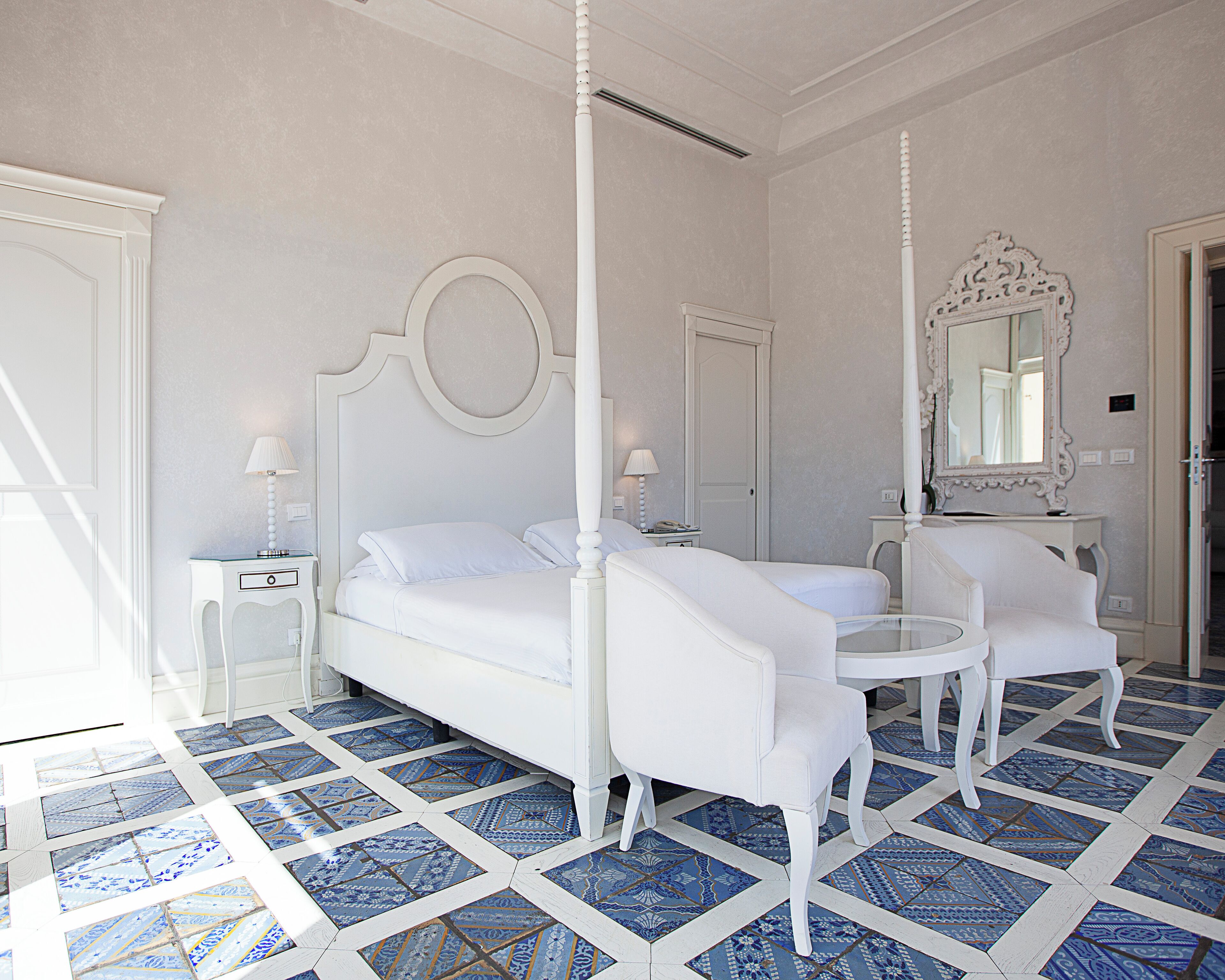 A four-poster bed with a round mirror above it, a small round table, two white chairs, and a patterned floor.