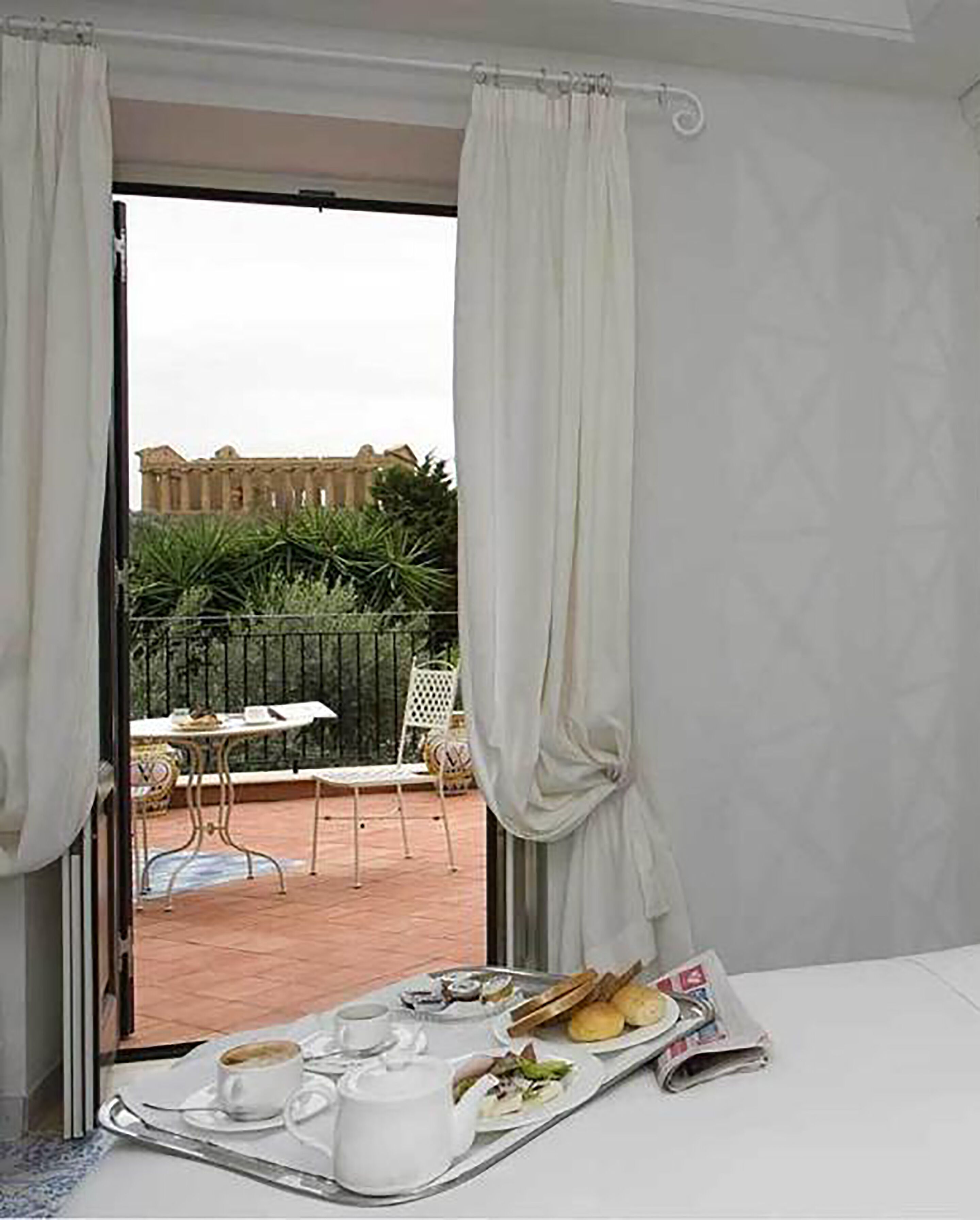 Ein Zimmer mit Balkon, einem Tisch mit Frühstücksgebäck und Blick auf ein Gebäude und Grünflächen.