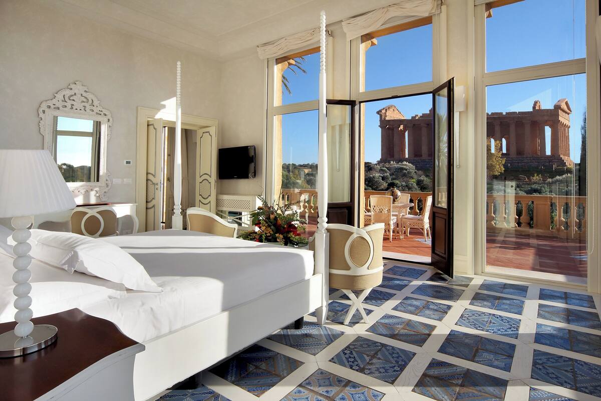 A hotel room with a large bed, a sitting area with a chair, a TV, and a balcony with a view of a historical building.