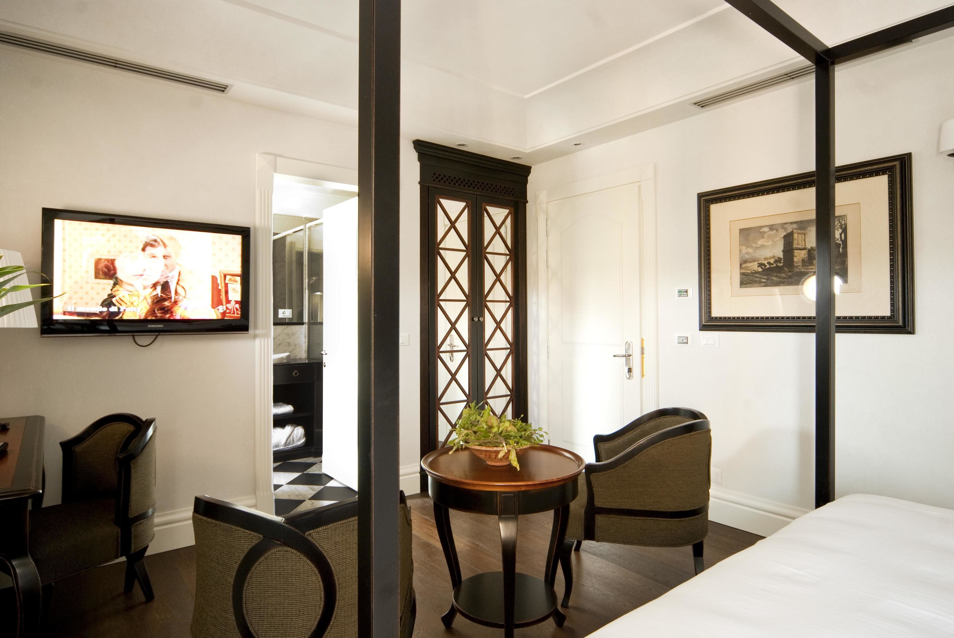 Une chambre d’hôtel comprenant un lit, une télévision, une table à manger avec des chaises, une plante en pot et une porte ornée d’un motif décoratif.