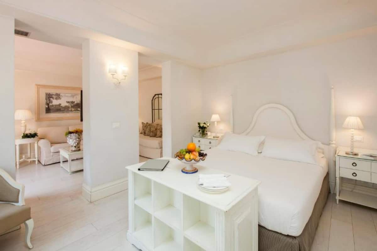 A white-themed bedroom with a large bed, bedside tables, a small white table with fruit, and a chair in the background.