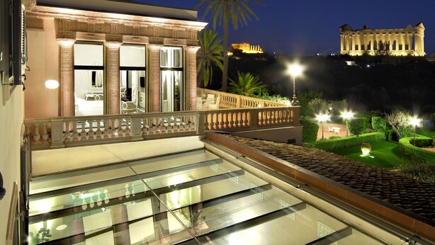 Ein Balkon mit Glasgeländern, der auf einen gut beleuchteten Garten und ein historisches Gebäude im Hintergrund blickt.