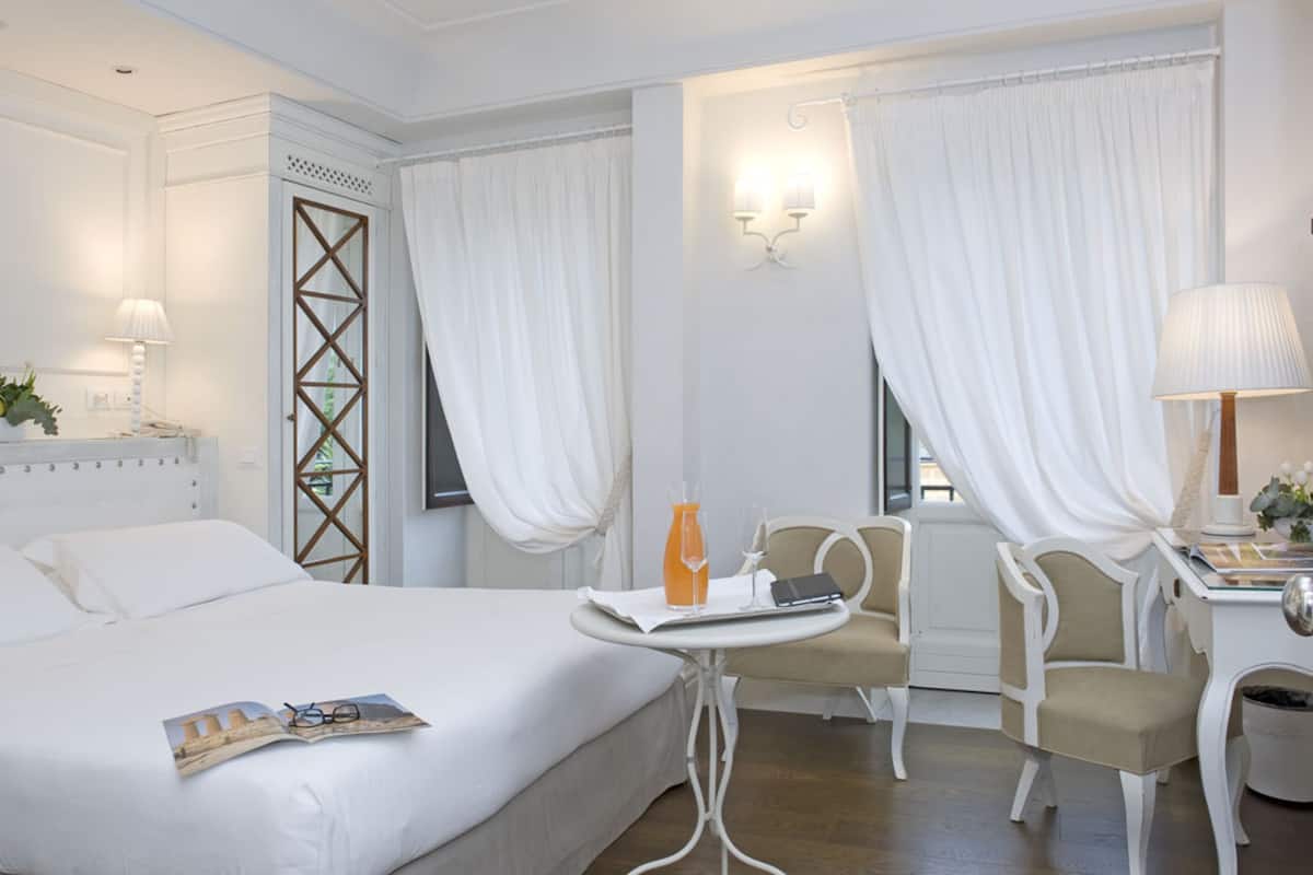 A hotel room with a bed, a small table with a drink, a chair, and a desk with a lamp.