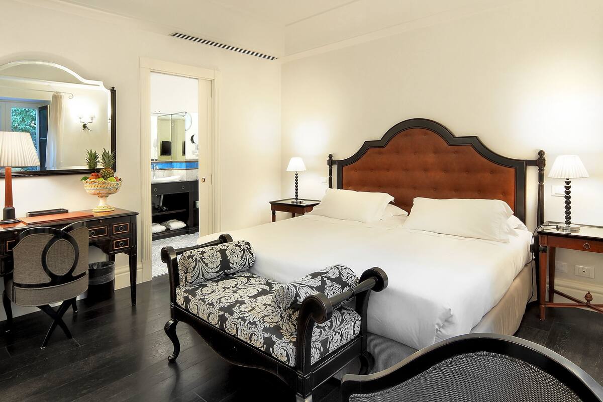A hotel room with a large bed, a desk with a chair, a mirror, and a nightstand with a lamp.