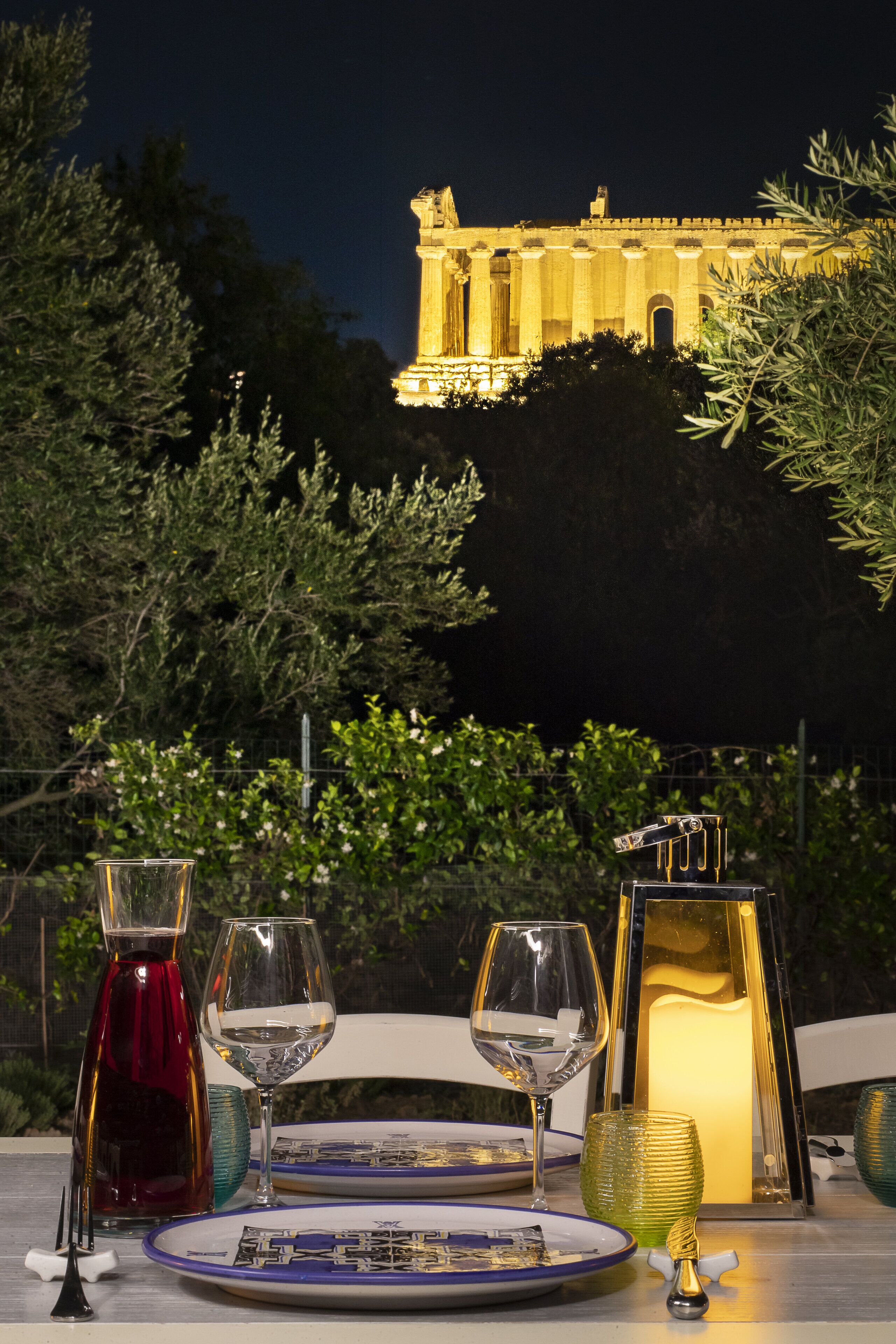 A table set for two with wine glasses, a candle, and a decorative glass pitcher. In the background, a building with classical architecture is illuminated at night.