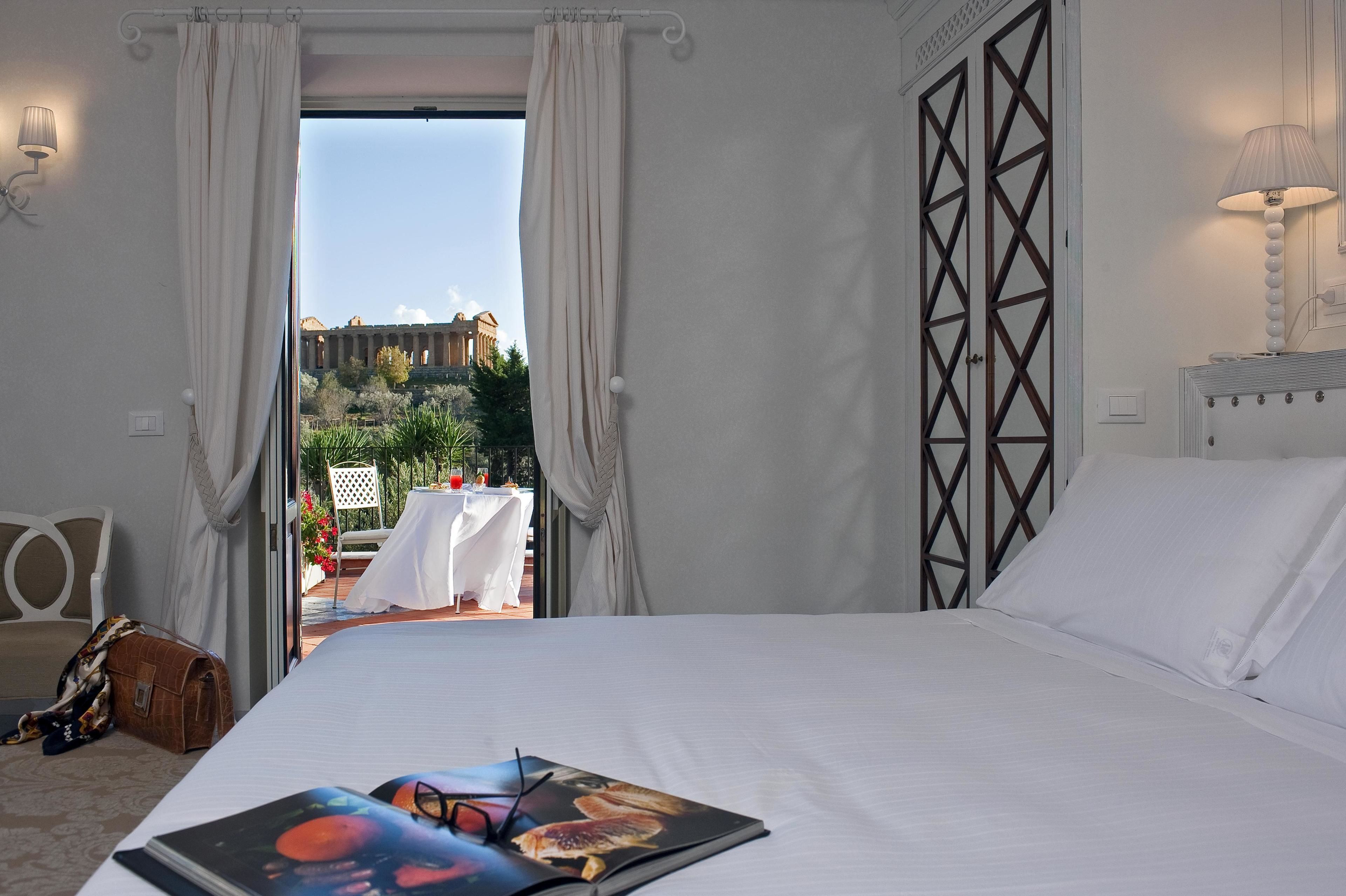 A hotel room with a bed, a view of the outdoors, a table with a white tablecloth, and a lamp.