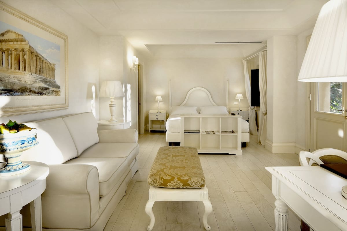 A hotel room with a bed, a white sofa, a bench, a nightstand, and a lamp.