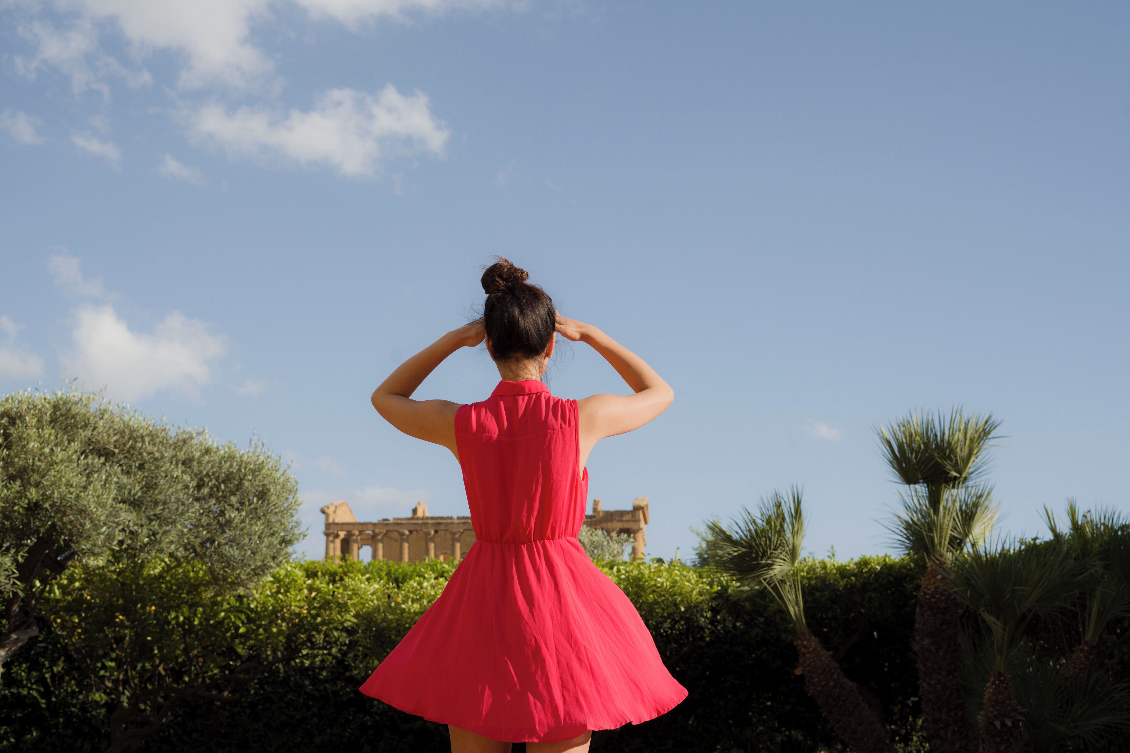 A woman in a red dress standing with her back to the camera, facing a historic building with columns and a clear blue sky above.