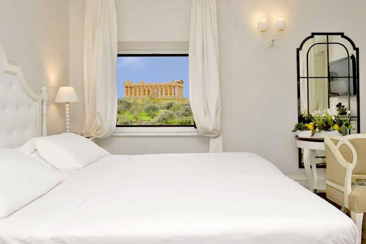 A bedroom with a bed, a nightstand, a lamp, a window with curtains, a mirror, and a view of the Parthenon.