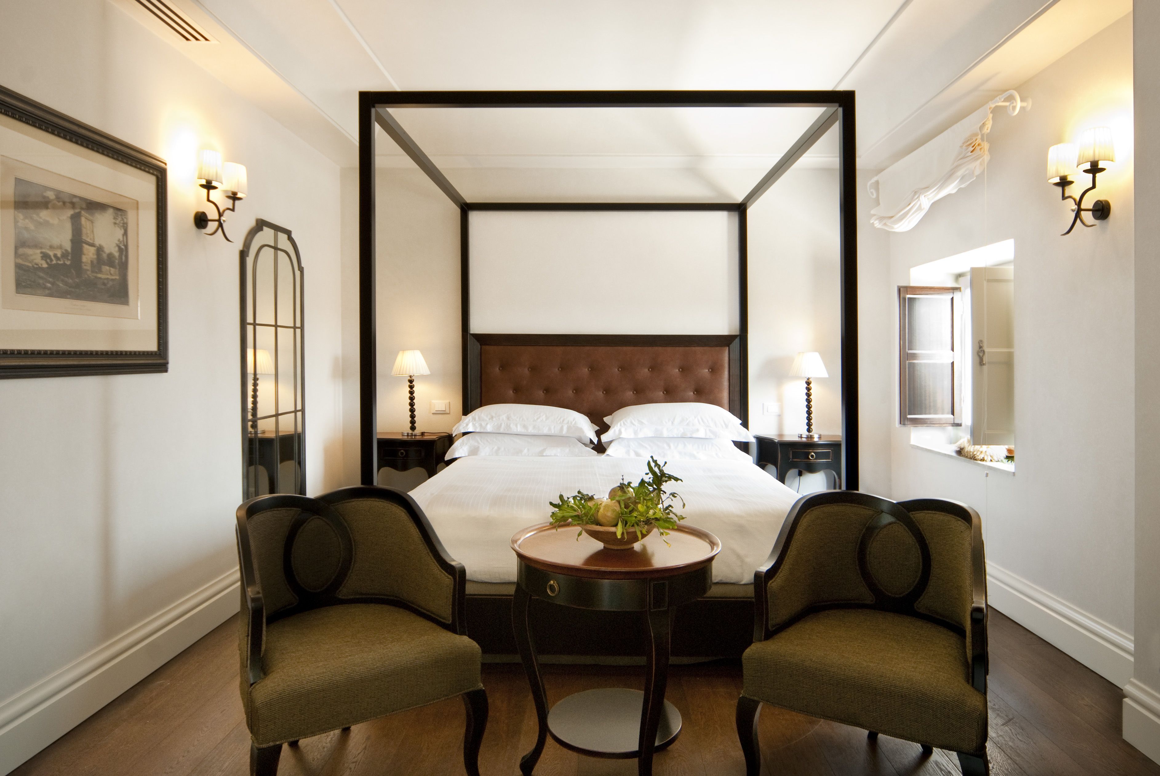 A four-poster bed with white linens, two upholstered chairs, a small round table with a plant, and wall sconces.