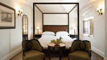 A four-poster bed with white linens, two upholstered chairs, a small round table with a plant, and wall sconces.