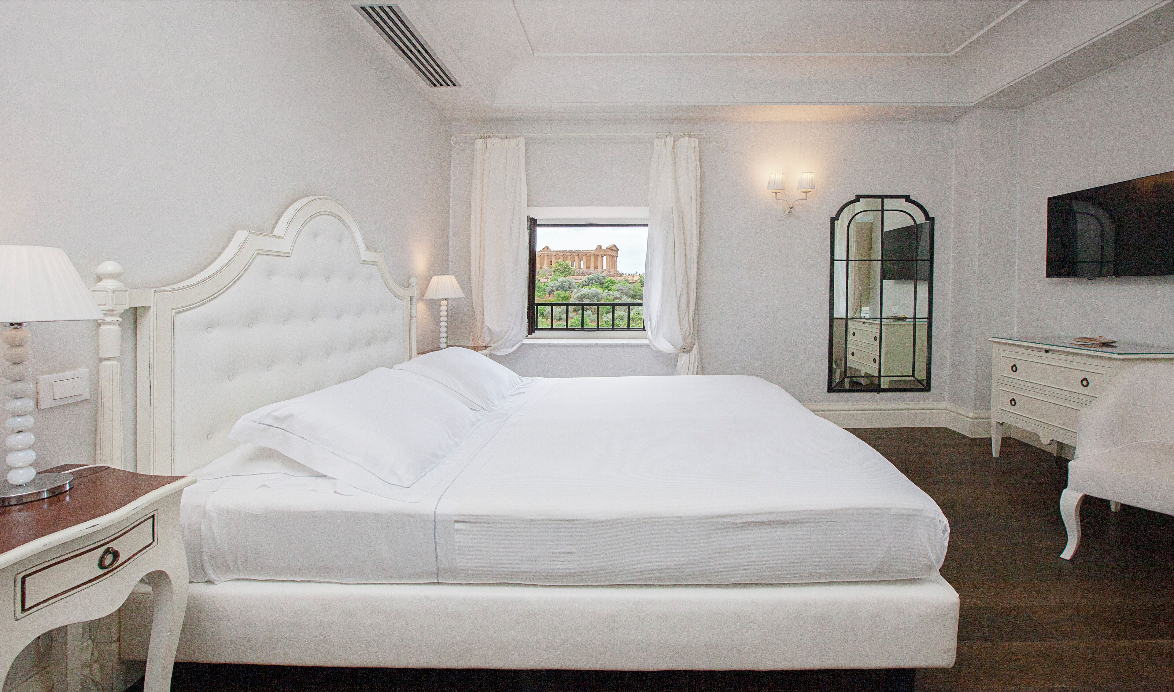 Habitación de hotel con una cama grande, una mesita de noche, un aparador, un espejo y vistas a un edificio histórico.