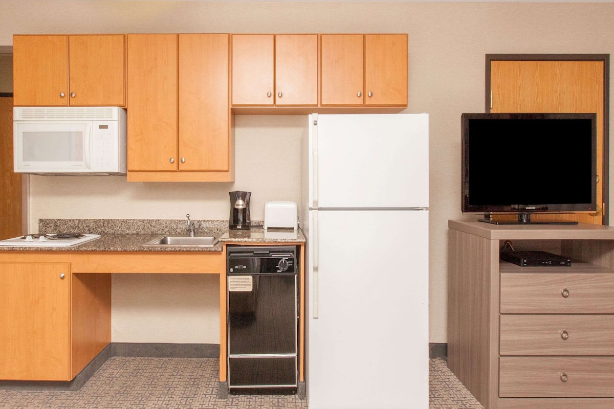 A compact kitchen with wooden cabinets, a microwave, a refrigerator, a dishwasher, and a television on a wooden stand.