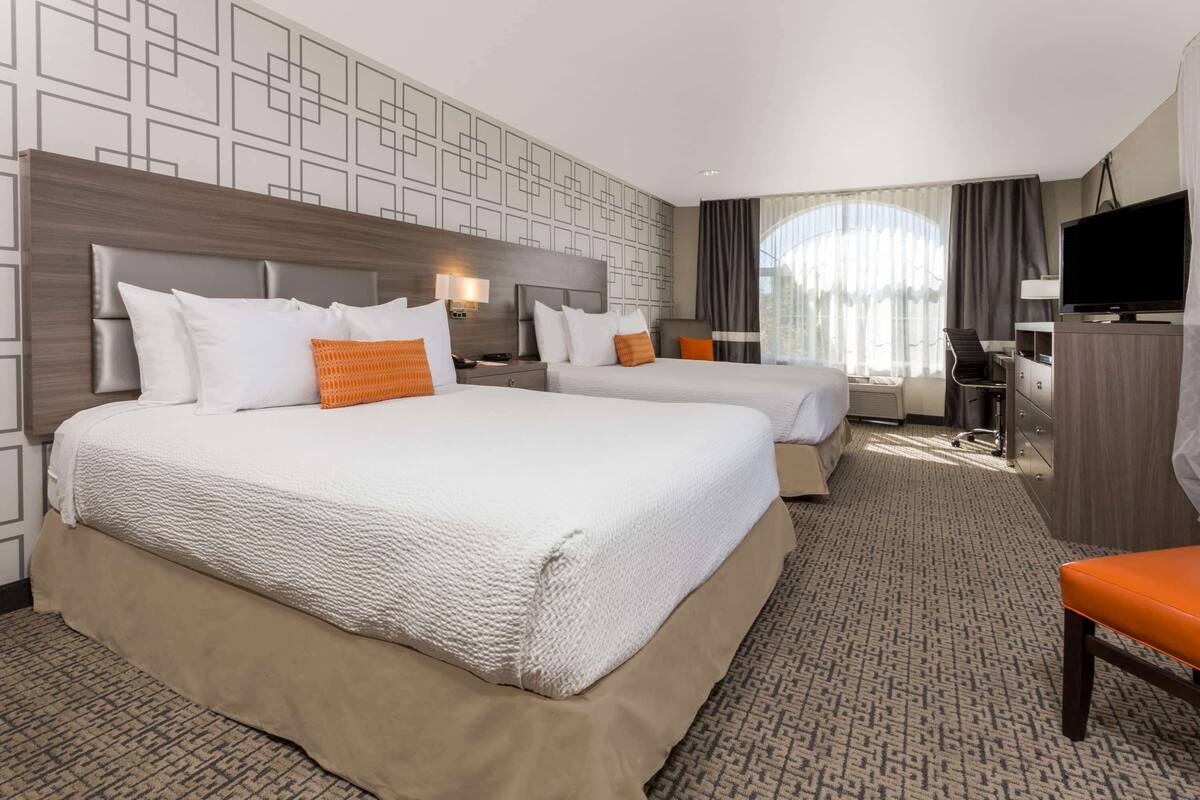 A hotel room with two beds, a desk, a television, and a large window with curtains.
