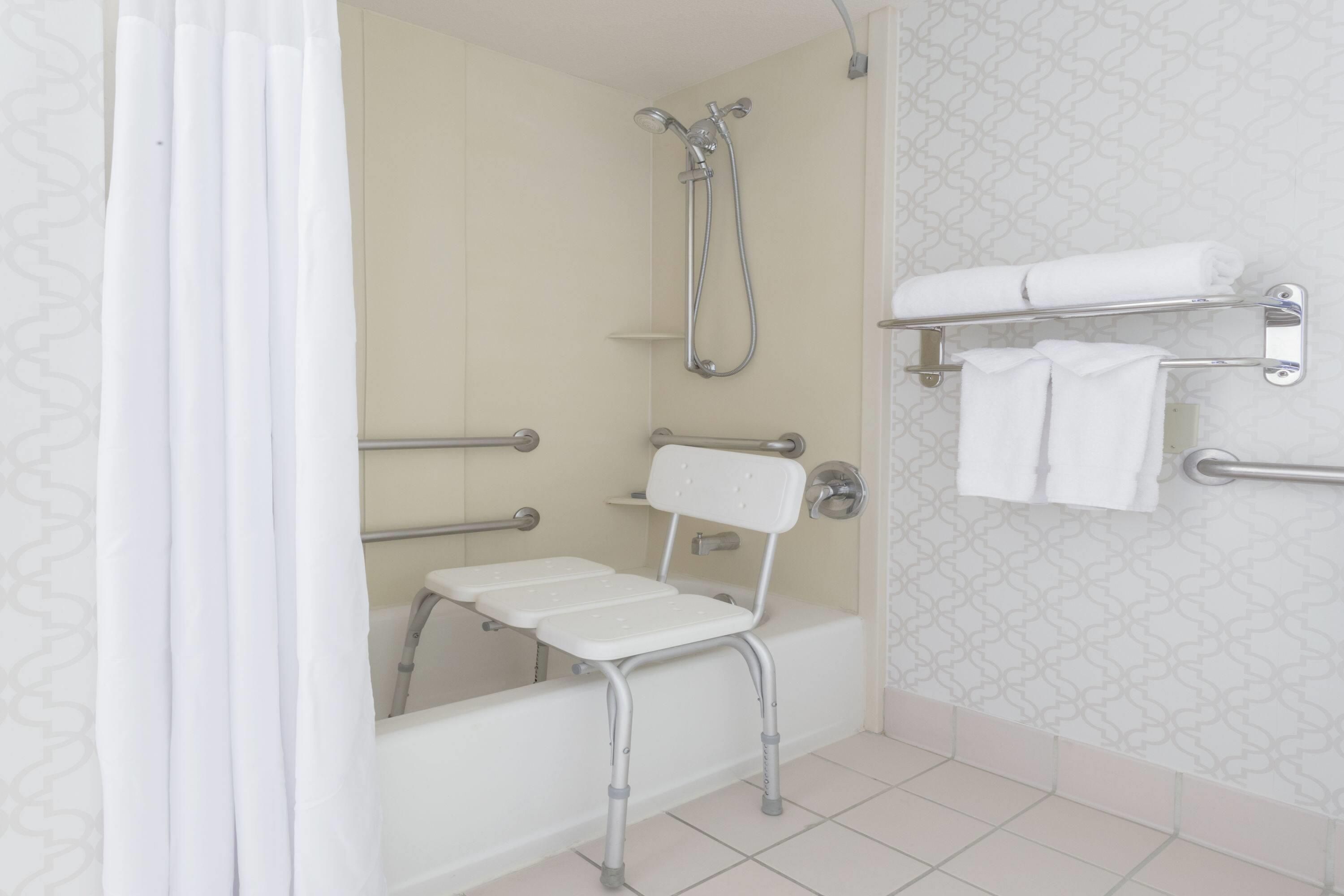A bathroom with a shower, grab bars, a white chair, and a towel rack.