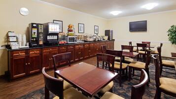 A hotel dining area with a buffet setup, wooden chairs, a clock, and a TV on the wall.