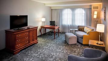 A hotel room with a flat-screen TV, wooden dresser, desk, office chair, sofa, ottoman, and a lamp.
