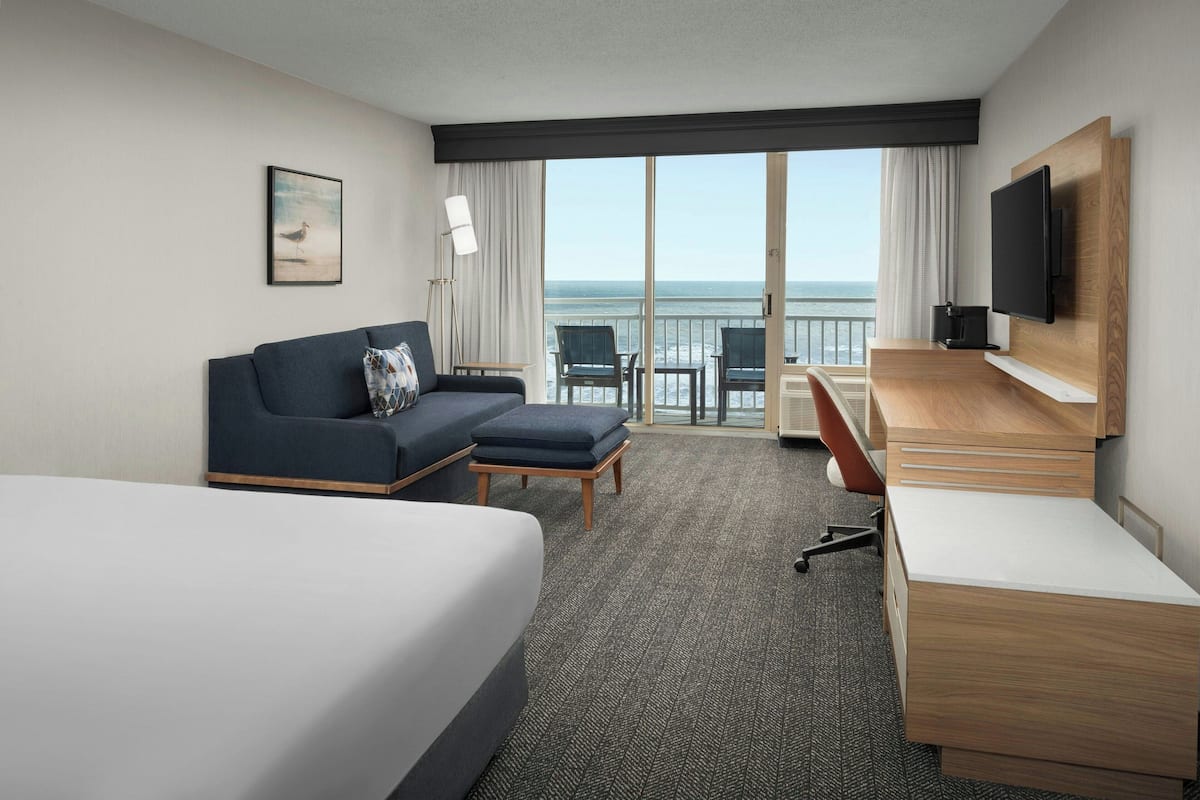 A hotel room with a bed, a sofa, a desk, and a TV. There is a view of the ocean through large windows.