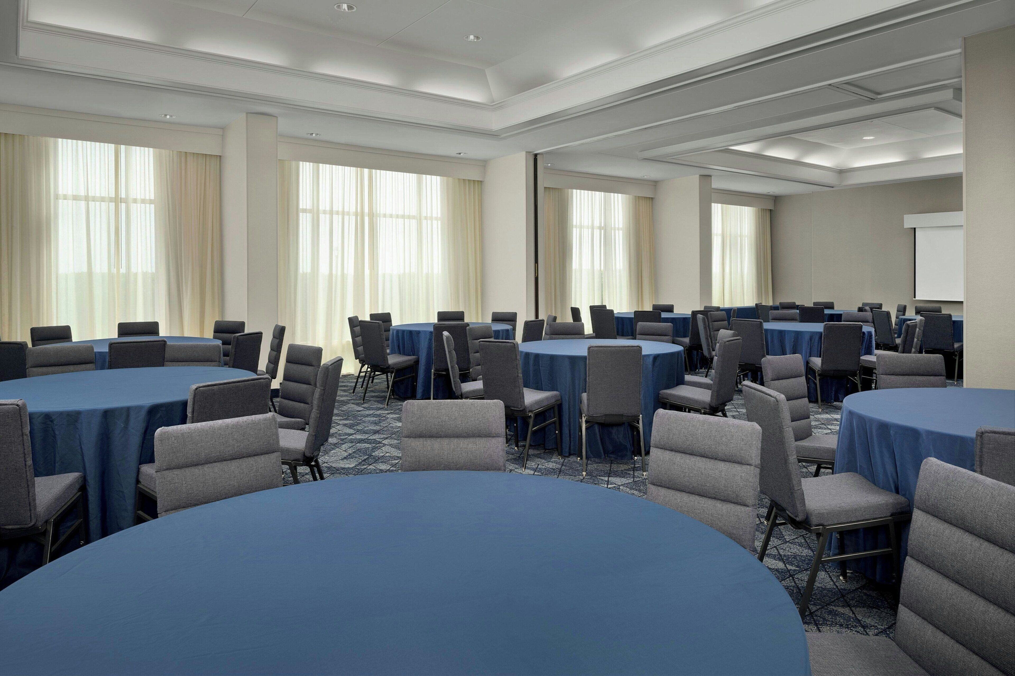 Un salón de conferencias con varias mesas redondas, cada una rodeada de sillas, y grandes ventanas con cortinas.