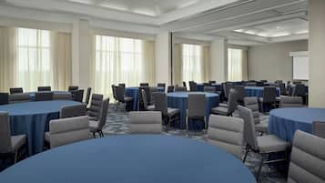 Un salón de conferencias con varias mesas redondas, cada una rodeada de sillas, y grandes ventanas con cortinas.