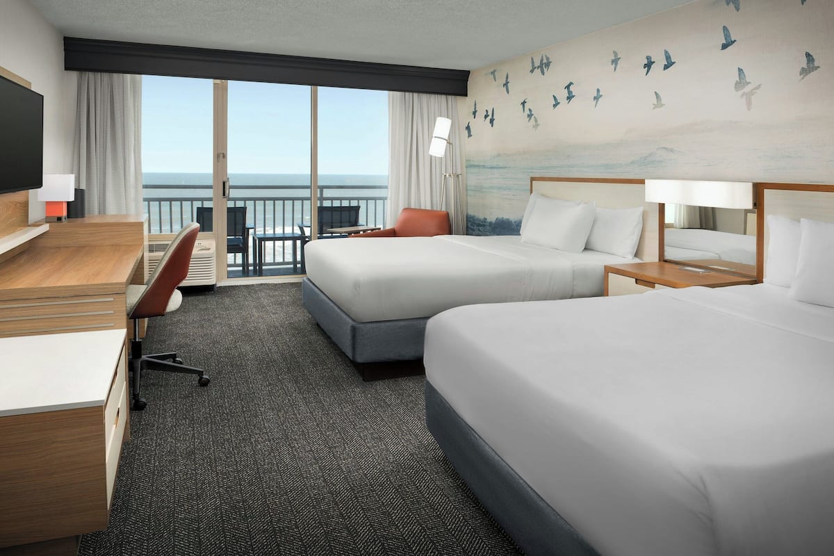 A hotel room with two beds, a desk, a chair, and a large window with a view of the ocean.