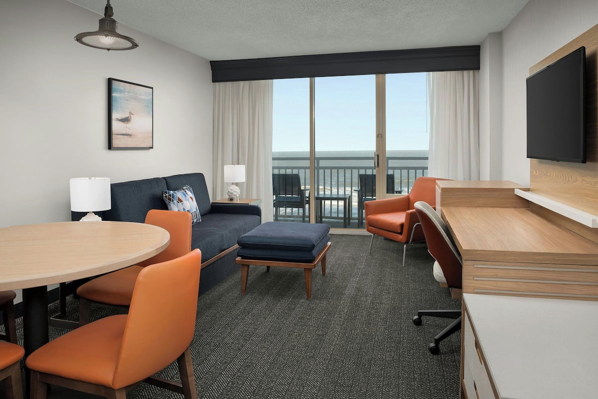 A hotel room with a balcony, a sofa, a desk, and a dining table.