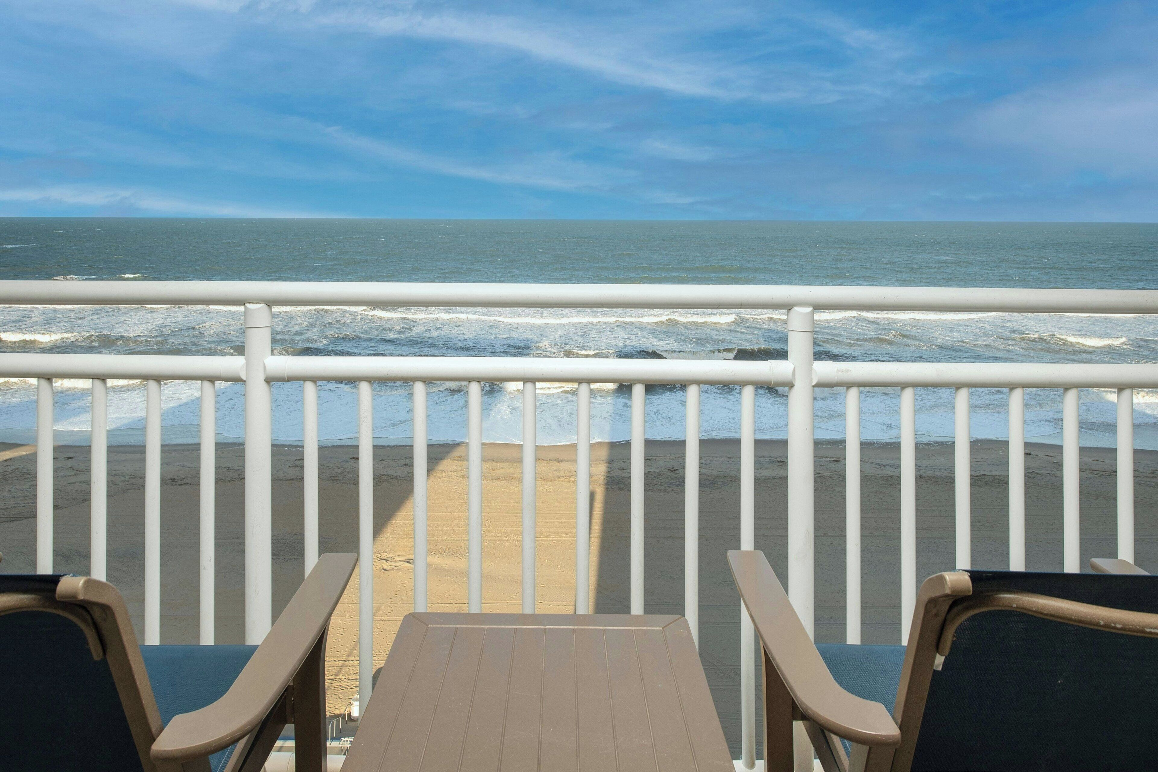 A deck with two chairs and a table, overlooking a beach with waves and a clear sky.