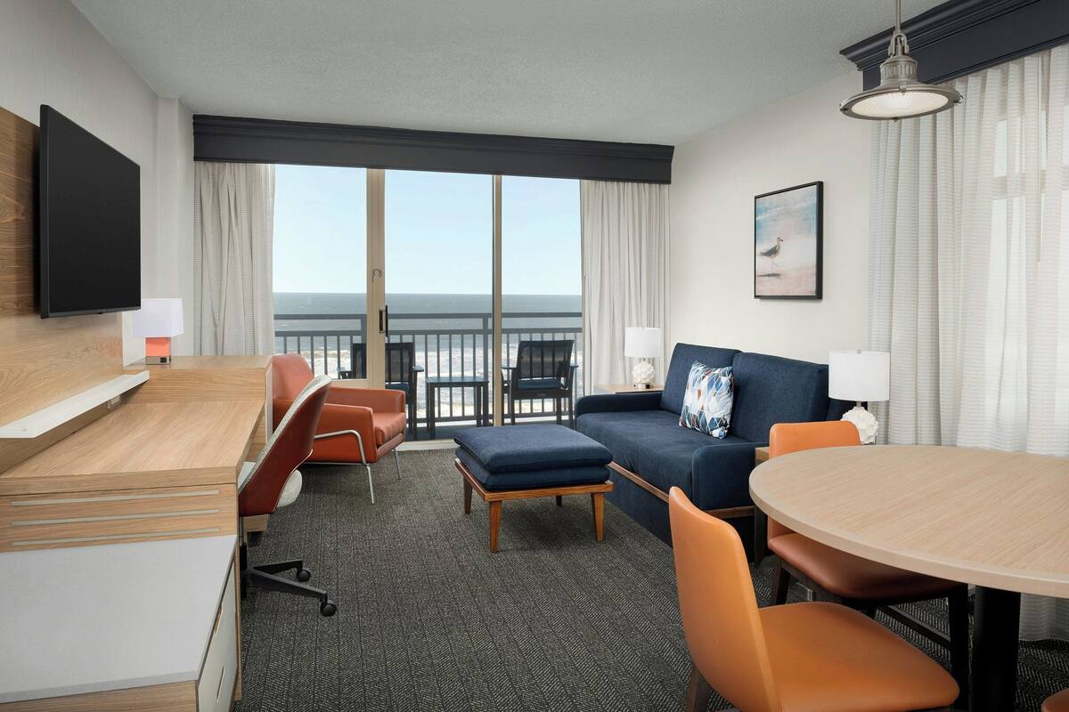 A hotel room with a desk, two chairs, a sofa, a round table, and a balcony with a view of the ocean.