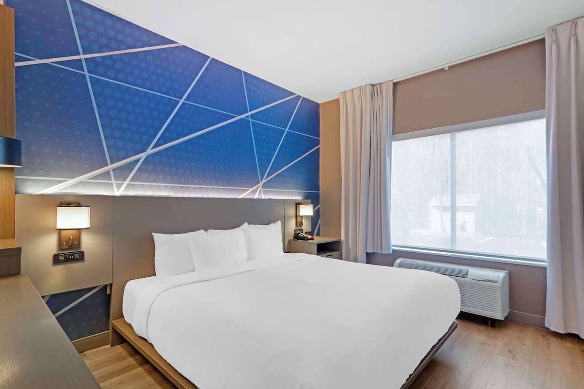 A modern hotel room with a large bed, bedside lamps, a nightstand, a window with curtains, and a geometric wall design.