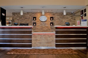 Una recepción con pared de piedra de fondo, un quiosco digital y un tapete de bienvenida.