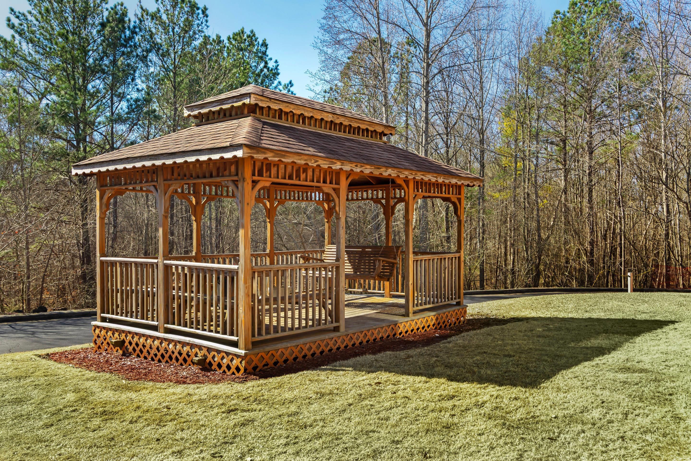 A wooden gazebo with a tiled roof, surrounded by a grassy area and trees.