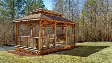 A wooden gazebo with a tiled roof, surrounded by a grassy area and trees.
