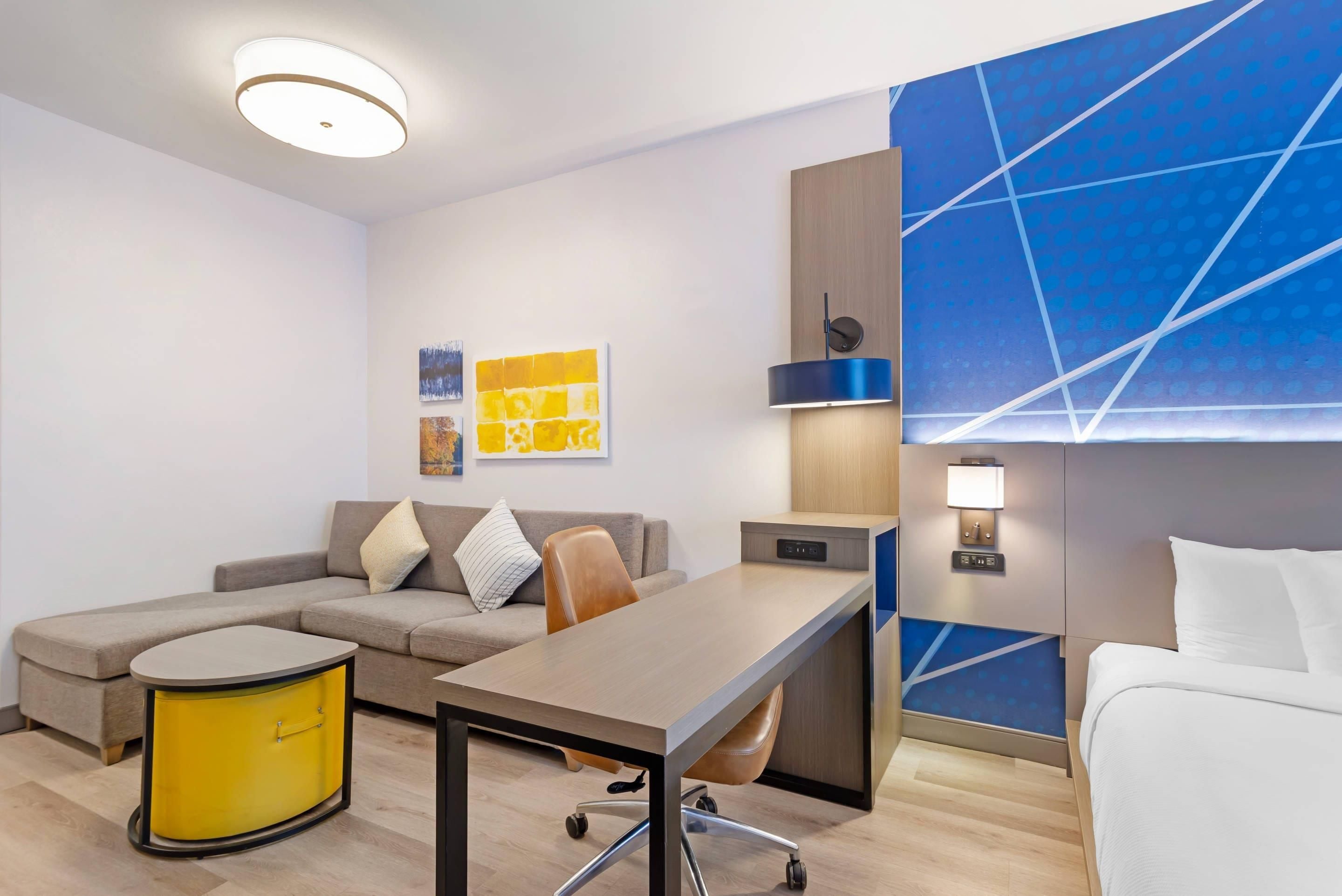 A modern hotel room with a sofa, a dining table, a yellow cabinet, a blue accent wall, and a ceiling light.