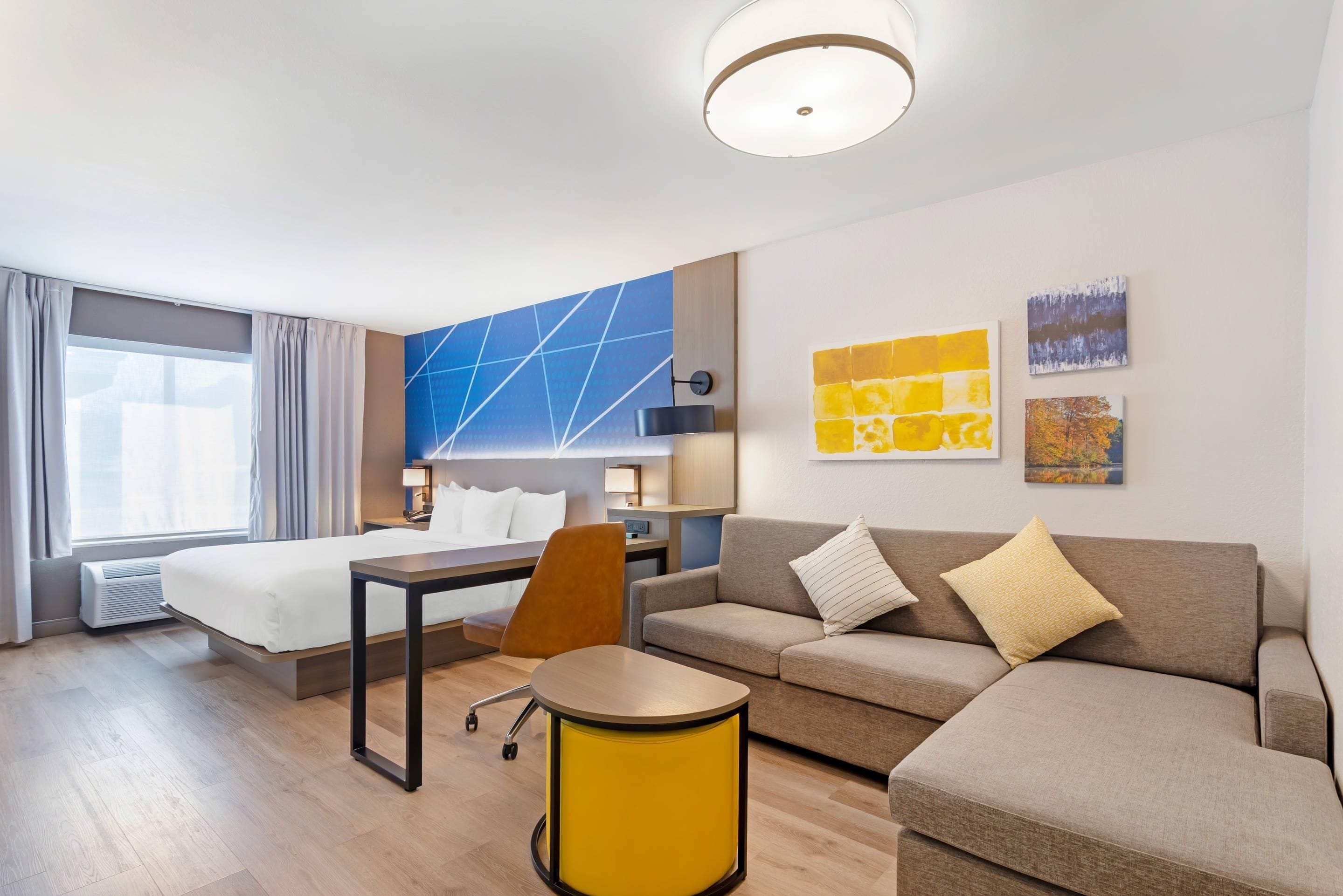 Habitación de hotel con cama, sofá, una mesita y una silla. Hay dos cuadros en la pared y una ventana con cortinas.