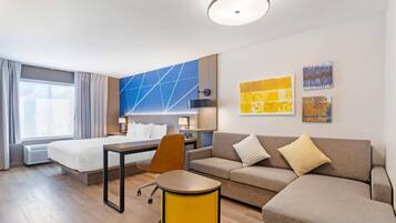 Habitación de hotel con cama, sofá, una mesita y una silla. Hay dos cuadros en la pared y una ventana con cortinas.