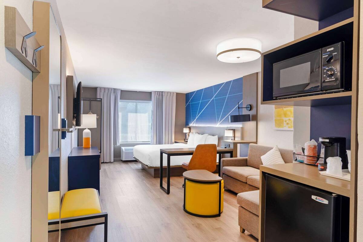 A hotel room with a bed, a desk with a computer, a chair, a sofa, a small table, a microwave, and a refrigerator.