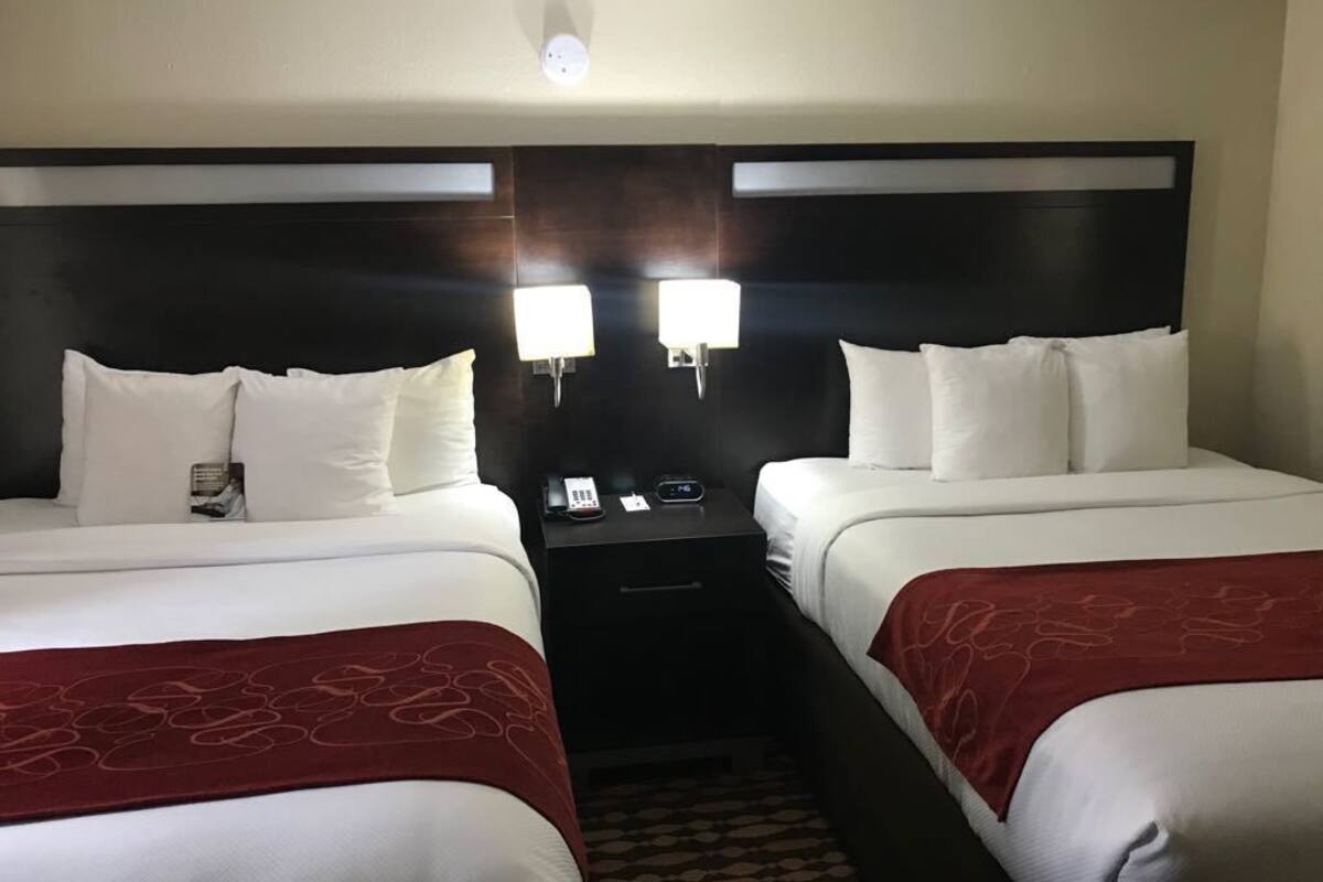 A hotel room with two beds, a nightstand with a phone and a remote, and wall-mounted lights.
