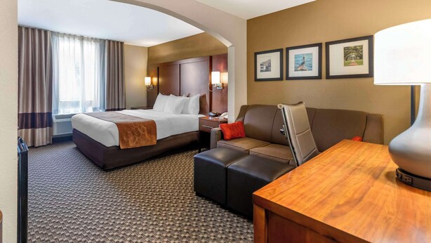A hotel room with a bed, a sofa, a desk, and a lamp. There are three framed pictures on the wall. - Comfort Suites Port Allen - Baton Rouge (Port Allen)