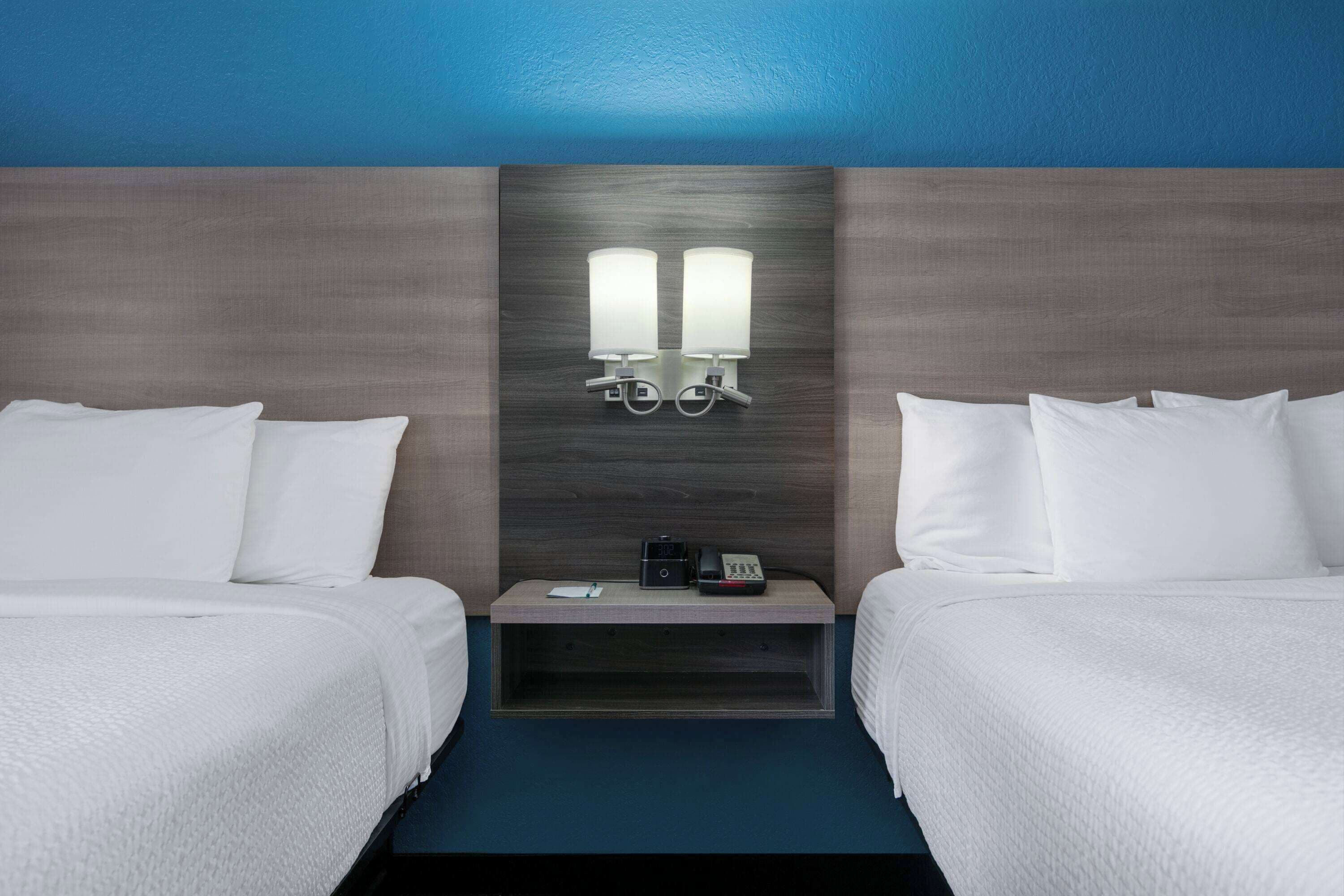 A hotel room with two beds, a nightstand, and a wall-mounted lamp.